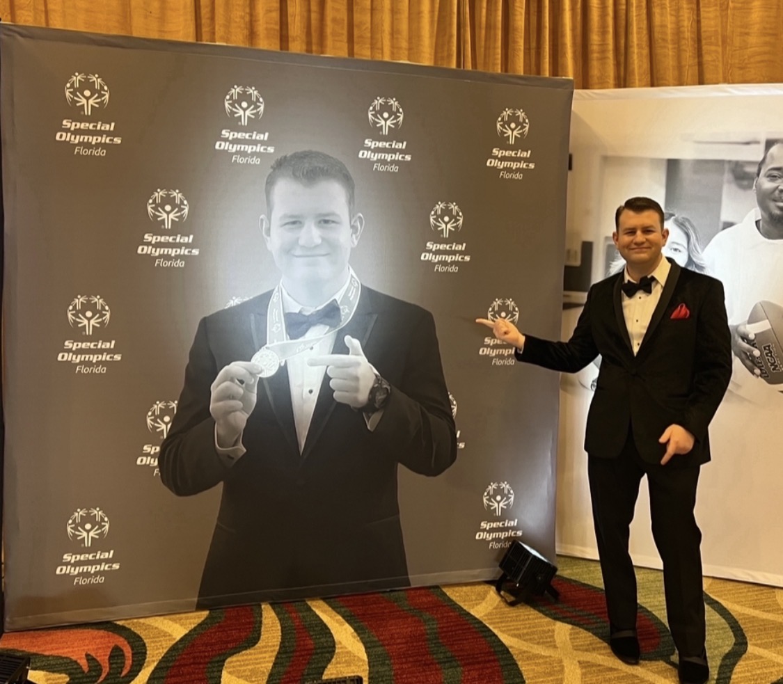 Stephen in a tuxedo beside his large Special Olympics Florida portrait backdrop